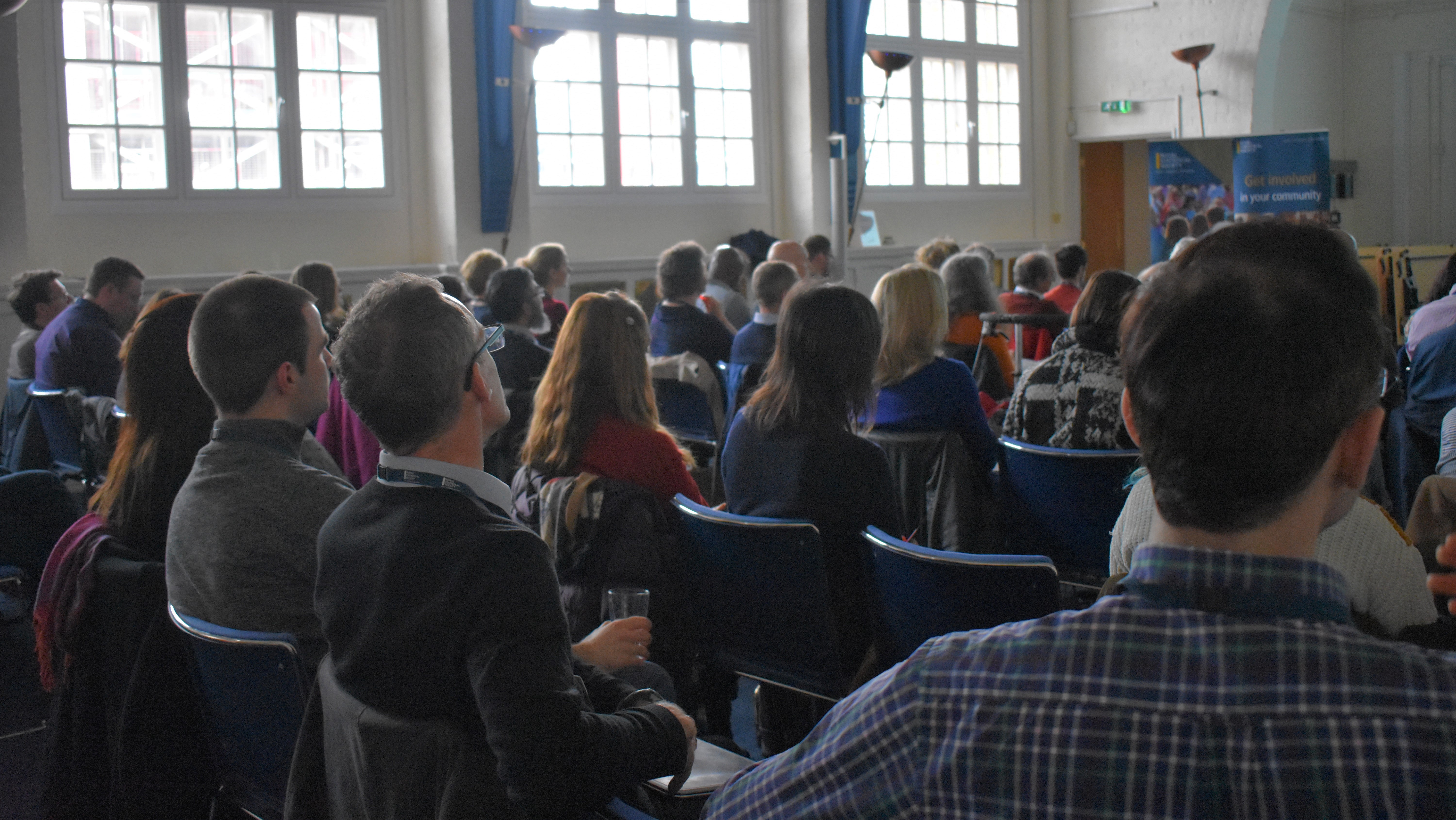 Back of an audience sitting on chairs looking in the direction of a speaker.
